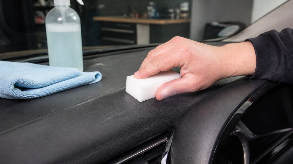 Close-up van iemand die met een licht vochtige melaminespons voorzichtig over een kunststof dashboard van een auto wrijft, met een microvezeldoek en een fles zeepsop op de achtergrond in een garage