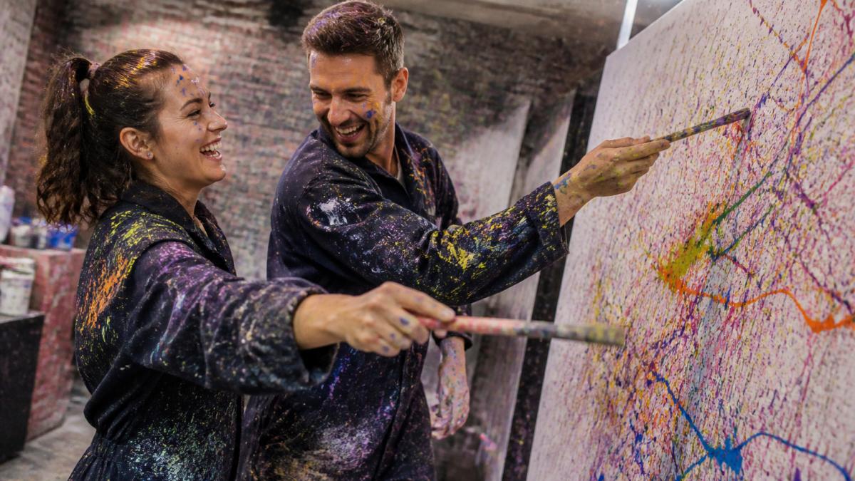 Ein Paar beim Schleuderbilder-Workshop, beide mit Pinseln in den Händen, bunte Farben fliegen auf eine große weiße Leinwand, beide lachen und sind von Farbspritzern bedeckt, helles Kunstlicht in der Werkstatt