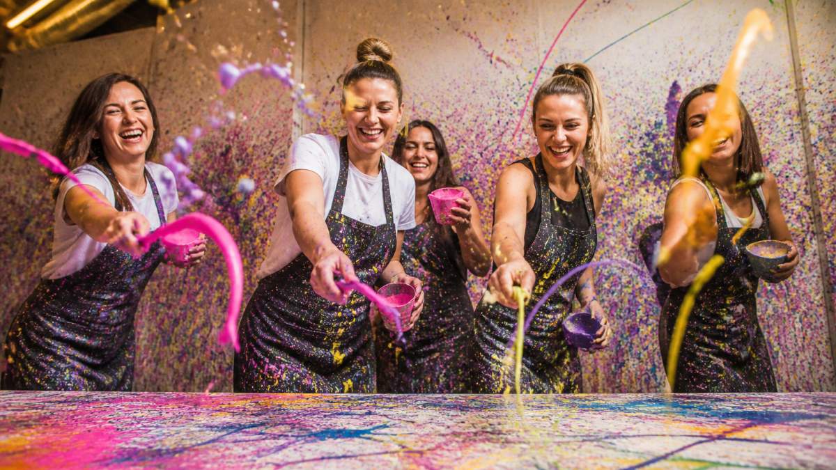 Eine fröhliche Gruppe von Freundinnen beim Action Painting Workshop, wie sie bunte Schleuderbilder erschaffen, dabei lachen und gemeinsam malen, mit bunten Farbtropfen in der Luft und hellen Lichtern
