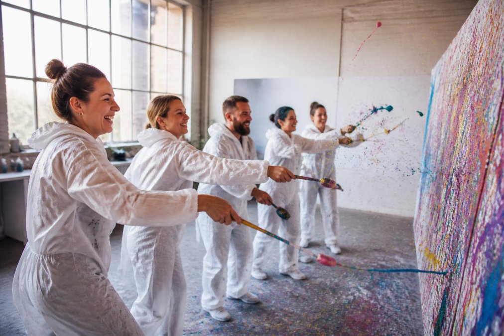 Eine Gruppe lachender Freunde in Malkitteln, die gemeinsam bunte Farbe auf eine große Leinwand schleudern, in einem hellen, kreativen Loft-Studio mit Tageslicht