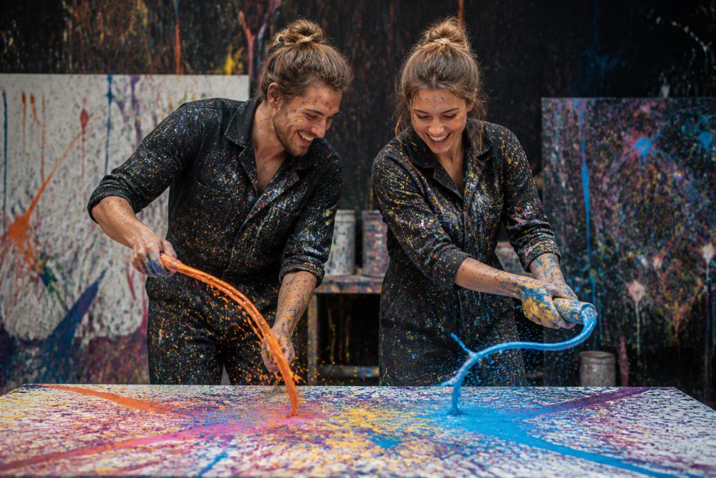 Ein Paar beim Action Painting Workshop, beide mit bunten Farbtropfen bespritzt, lachend und konzentriert bei der gemeinsamen Erstellung ihrer Schleuderbilder, lebendige Farben und dynamische Bewegung im Hintergrund