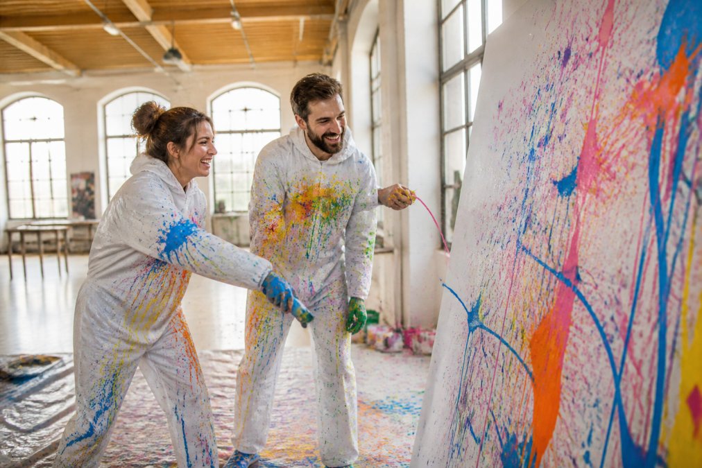 Ein Paar in weißer Schutzkleidung, das gemeinsam Farbe auf eine große Leinwand schleudert, beide lachen und sind farbenfroh bespritzt, in einem hellen Kunstatelier mit hohen Decken und Holzboden