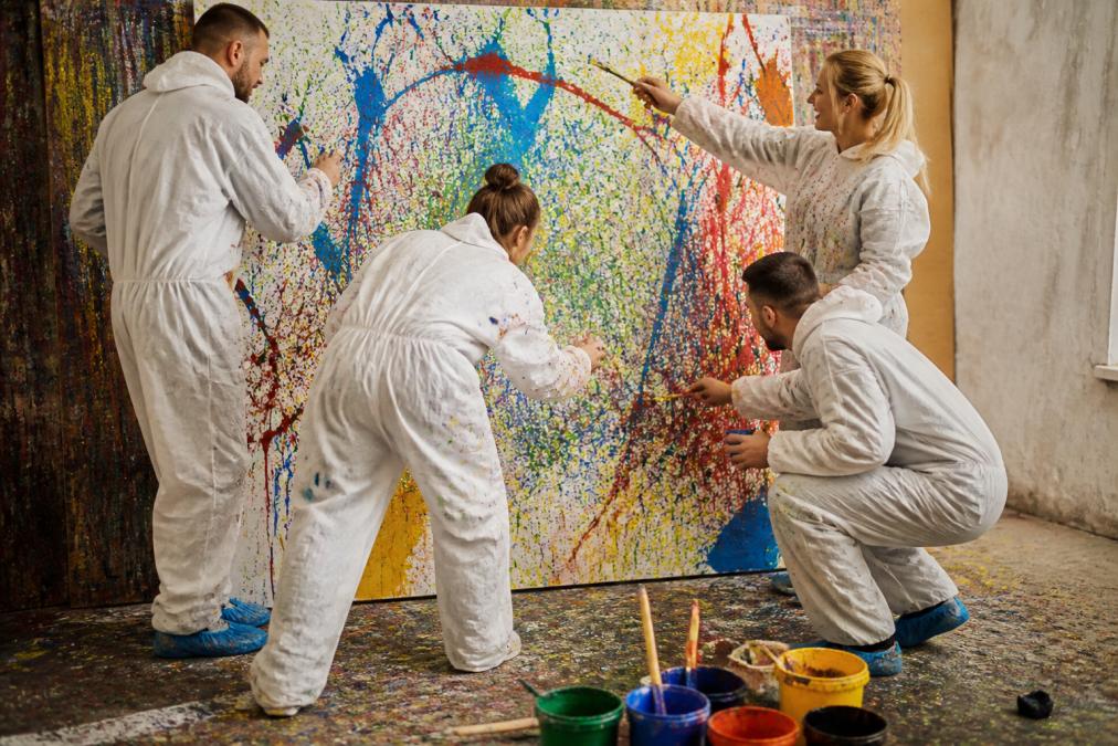 Ein Team von vier Mitarbeitern in Schutzanzügen, die gemeinsam an einer großformatigen Leinwand arbeiten, mit bunten Farben und Pinseln in einer inspirierenden Atelier-Atmosphäre