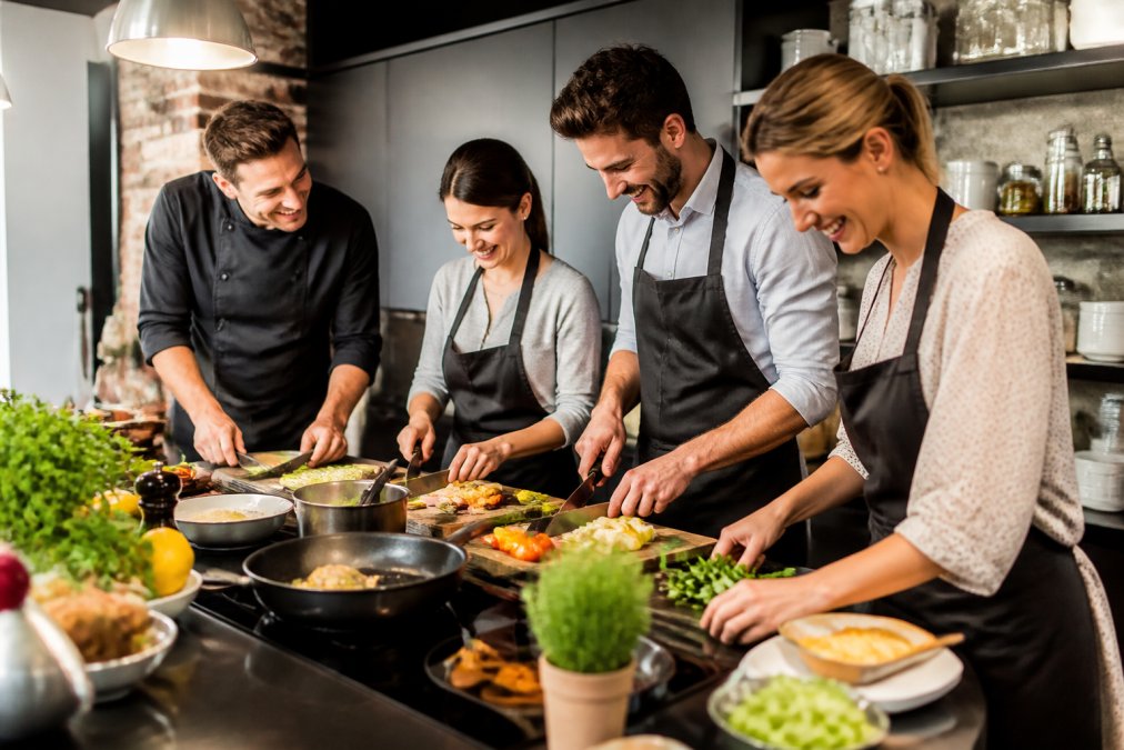 Ein Team beim gemeinsamen Zubereiten eines Menüs in einer modernen Küche mit einem Privatkoch, lachend und konzentriert bei der Arbeit