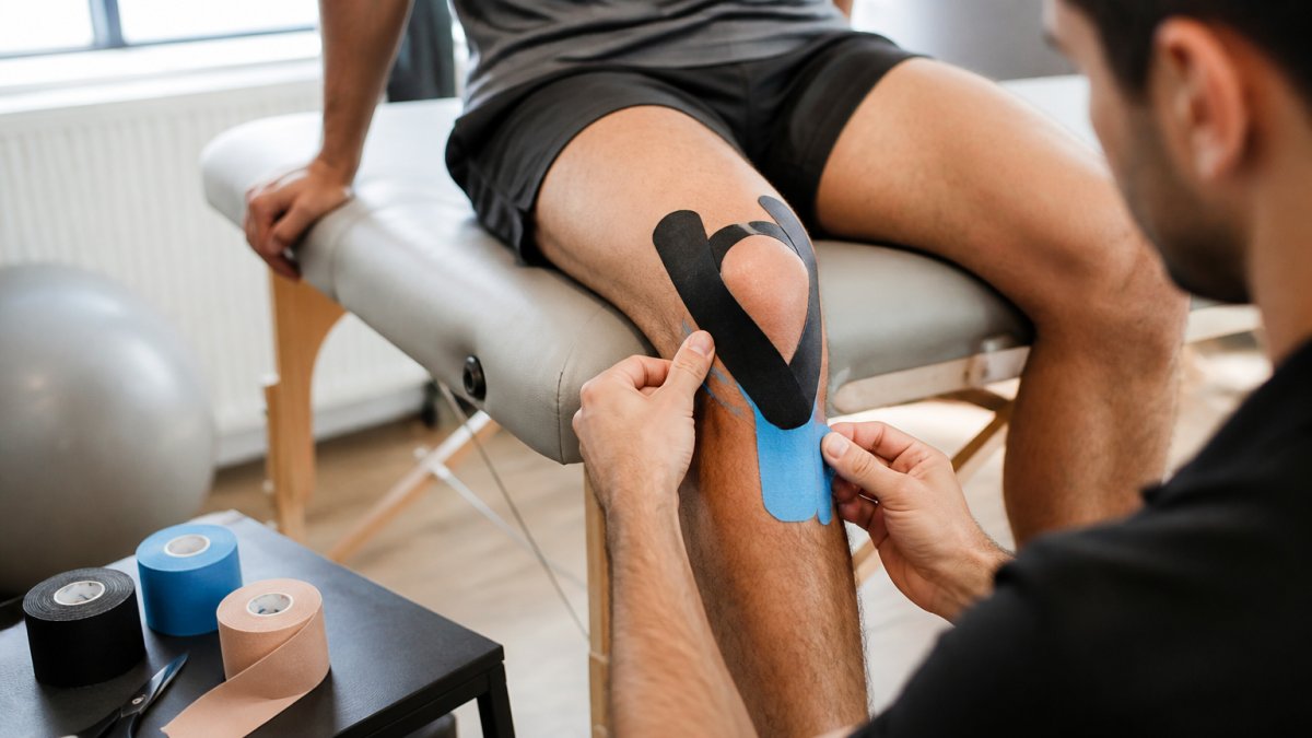 Professional demonstrating kt tape application technique on knee with proper hand positioning and tension during taping in clinical setting with natural lighting