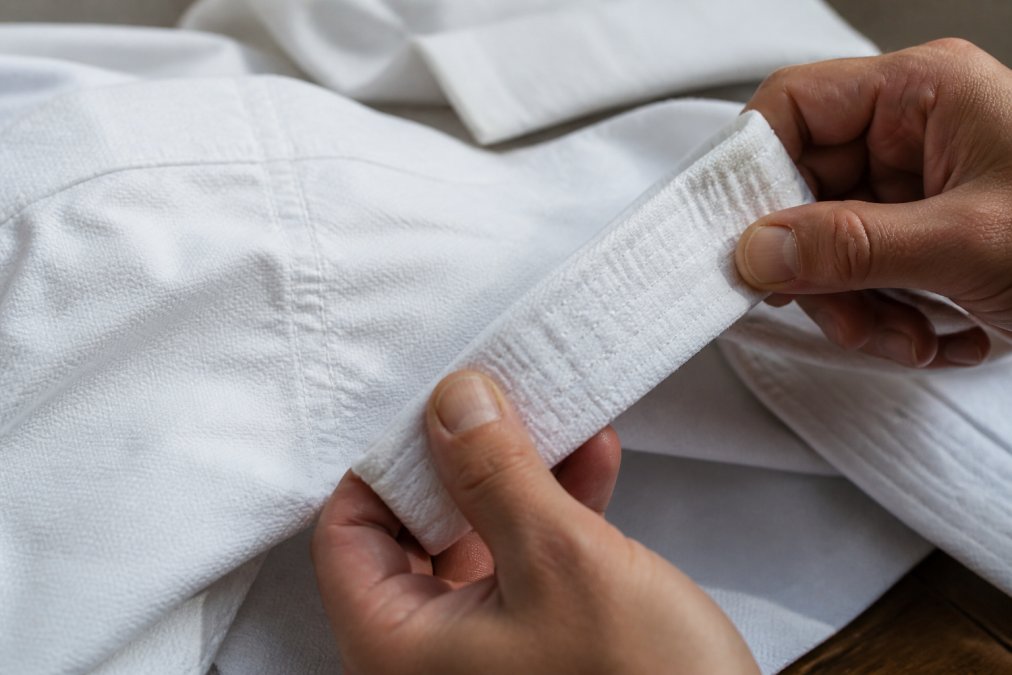 Close-up of hands examining high-quality martial arts uniform fabric showing detailed 55/45 cotton-polyester weave and reinforced double stitching at seams, with natural studio lighting