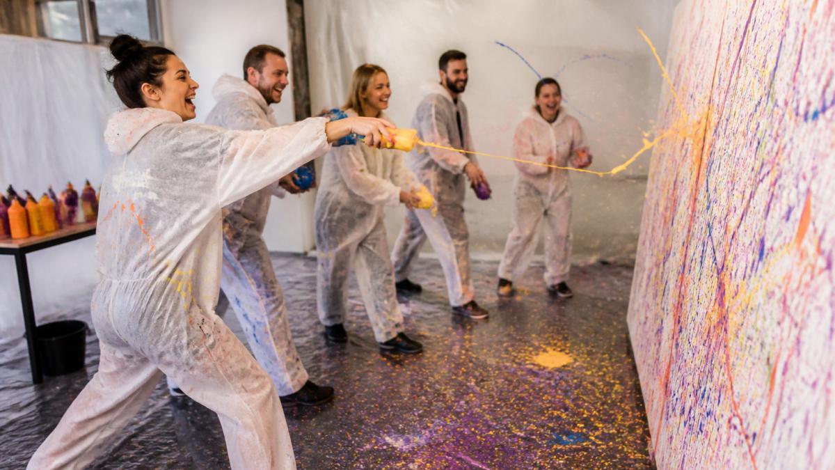 Eine Gruppe von Erwachsenen in Schutzkleidung schleudert Farbe auf eine große Leinwand in einem hellen Atelier, lachend und voller Energie, Farbspritzer auf dem Boden und den Kleidern