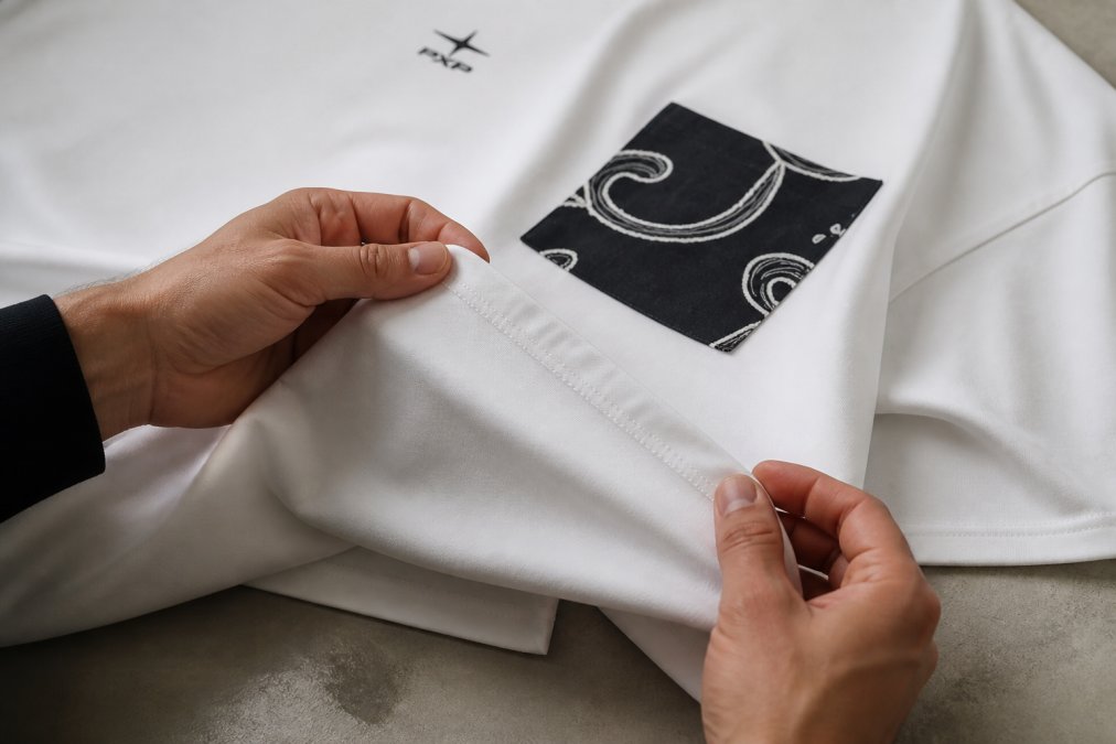 Close-up van handen die de stiksels langs de zijnaad van een oversized streetwear T-shirt controleren, met duidelijk zicht op de regelmatige steekpatroon en naadkwaliteit in natuurlijk licht