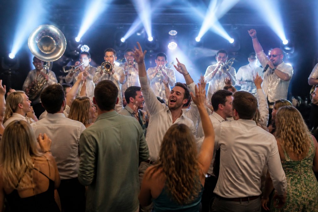 Live brassband op podium in volle interactie met dansend en enthousiast publiek, energieke sfeer met zichtbare emotie en beweging, fel podiumlicht en springende gasten