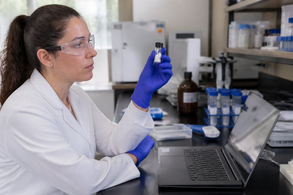 A laboratory researcher in a white coat examining a vial of lyophilized peptide powder next to a laptop displaying a certificate of analysis document