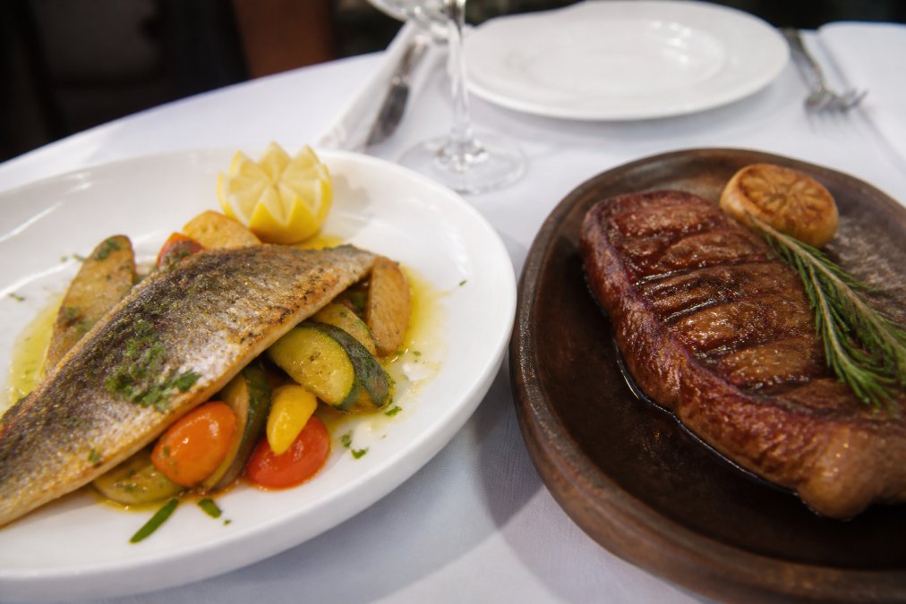Close-up of a beautifully plated Mediterranean seafood dish featuring pan-seared branzino with fresh herbs, lemon-infused olive oil drizzle, and roasted seasonal vegetables arranged artfully on a white plate, alongside a perfectly seared Argentine beef steak with crust on warm charred ceramic