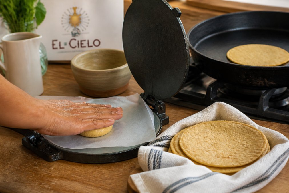 Hands pressing corn masa dough in a tortilla press beside a stack of freshly cooked corn tortillas in a tea towel
