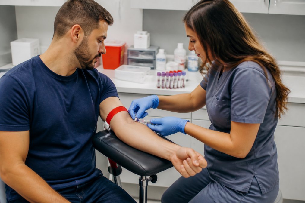 Healthcare professional drawing blood from a patient's arm during a lab visit, with collection tubes and medical supplies visible on the counter, clinical setting with professional lighting