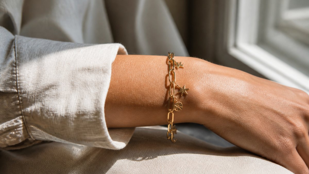 Close-up of a woman's wrist wearing a gold-plated charm bracelet with a connect link, styled with a linen sleeve and soft natural window light