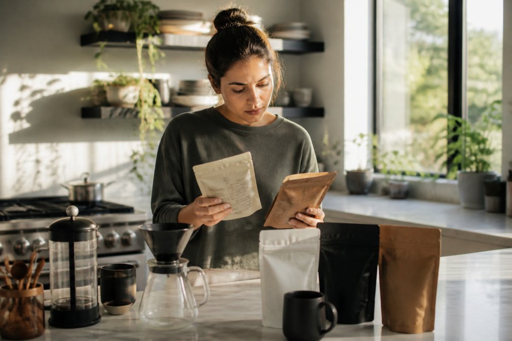 Person at a kitchen counter reviewing several coffee bags and brewing equipment with morning sunlight streaming through a window