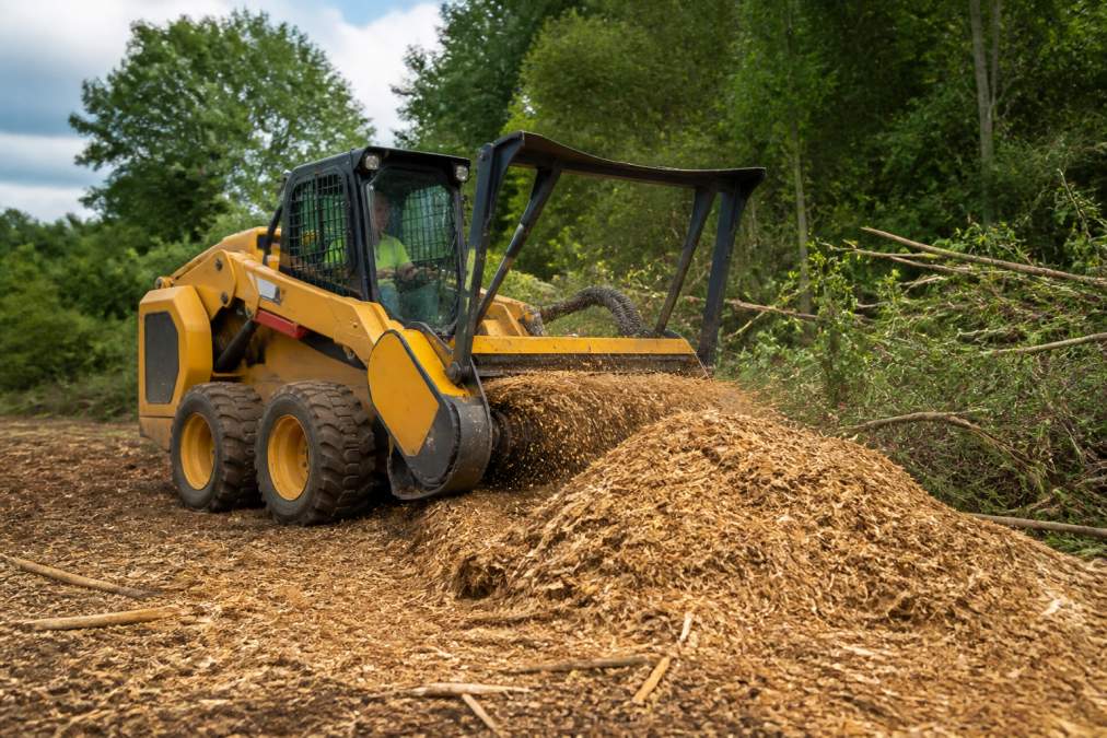 A forestry mulching machine with a wheeled base grinding brush and small trees into wood chips on a rural property, with an operator in the cab and a pile of mulch forming behind it