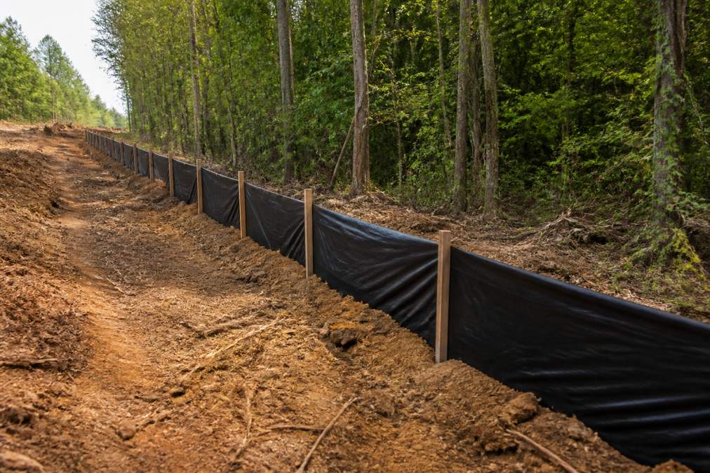 A silt fence installed along the downslope edge of a freshly cleared wooded property, black fabric staked into a shallow trench with exposed soil behind it and green trees in the background