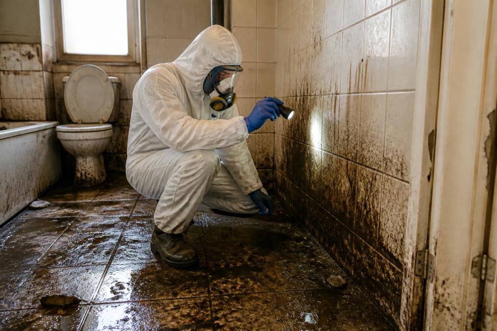 A professional technician in full PPE including white protective coverall, nitrile gloves, and respirator face mask crouching to inspect a flooded ground-floor bathroom with visible sewage contamination on tiles and lower wall surfaces under harsh overhead lighting