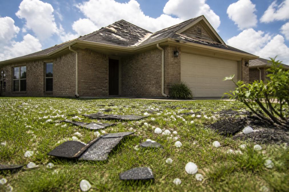 A Texas rental home with a damaged roof after a severe hailstorm, scattered shingles on the lawn, bright afternoon sun with clouds in the background