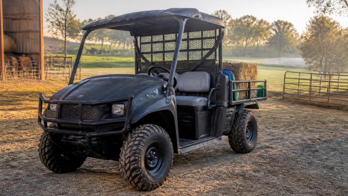 Landmaster UTV parked on a working farm with open fields, ranch equipment visible in background, showing the vehicle's all-steel frame and cargo bed loaded with farm supplies, morning sunlight across the property
