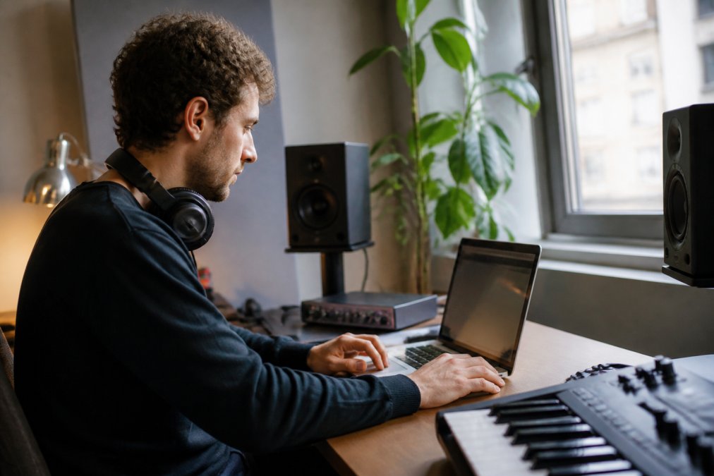 Independent music producer at a laptop reviewing distribution platform dashboard with headphones around neck and music production equipment visible in modern home studio with natural window lighting
