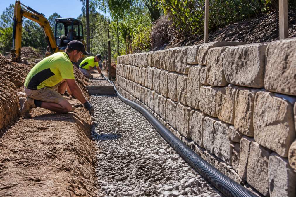 Professional landscape construction team installing stone retaining wall with proper drainage behind, showing excavated foundation and gravel layer, afternoon sunlight
