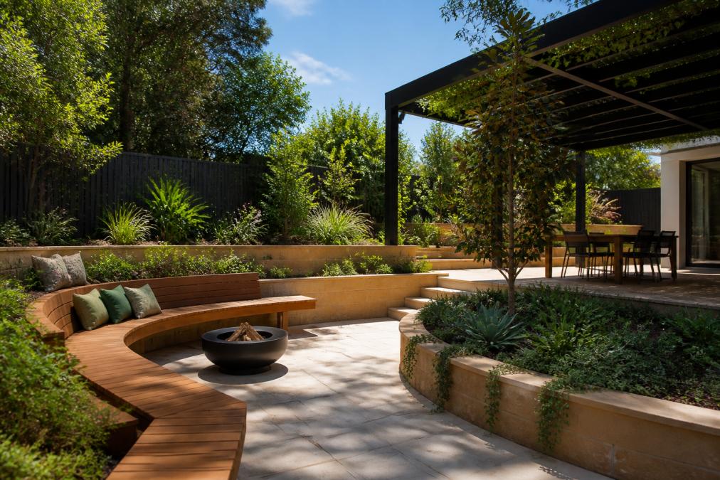 Contemporary residential garden showcasing curved paved entertaining area with timber seating, native plantings in tiered beds, and pergola overhead providing dappled shade on a sunny afternoon