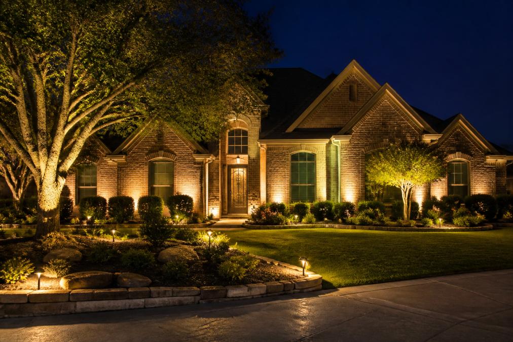 Front exterior of a residential home at night with professionally installed landscape lighting highlighting architectural features and uplighting on mature trees, showing warm color temperature with balanced brightness and layered illumination creating visual depth