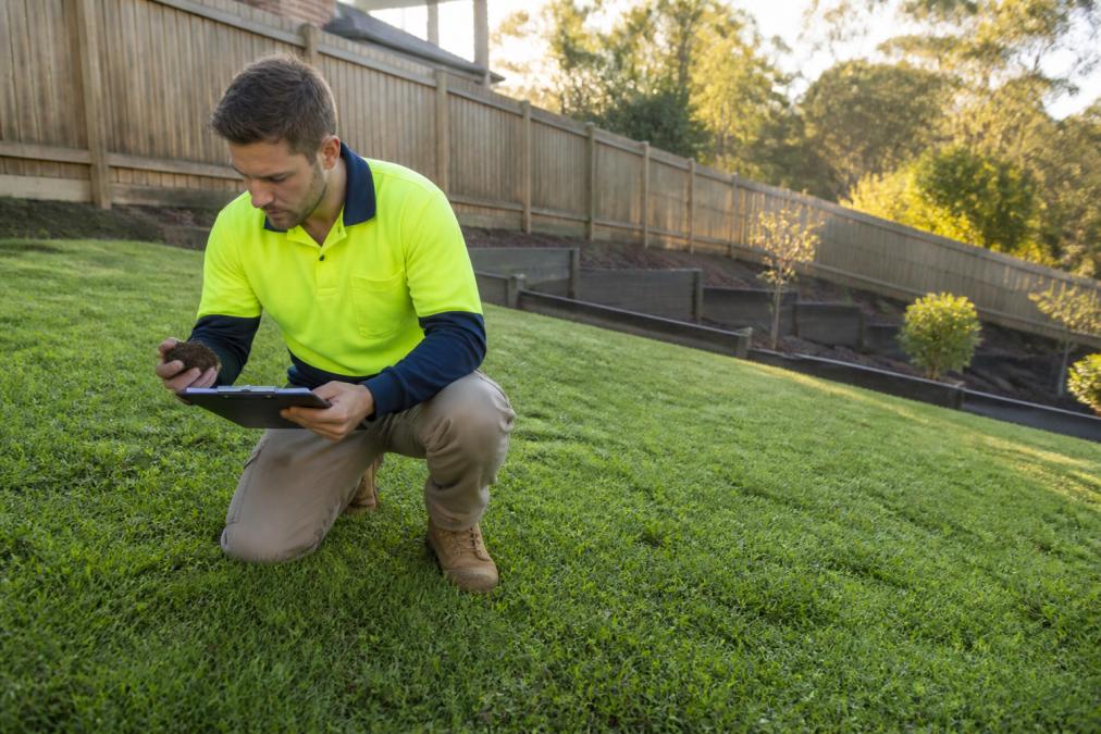 A landscaper Penrith professional examines soil in a residential backyard with a clipboard in hand.