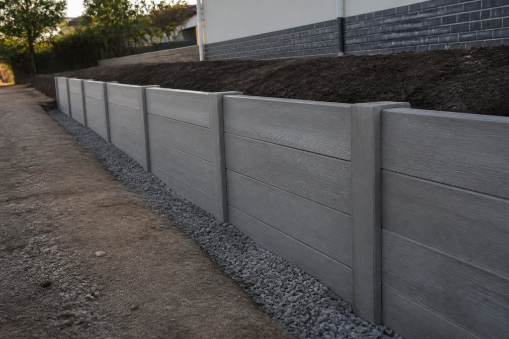 A newly constructed concrete sleeper retaining wall on a residential new build site, with clean drainage aggregate visible at the base and freshly laid soil behind it, late afternoon light