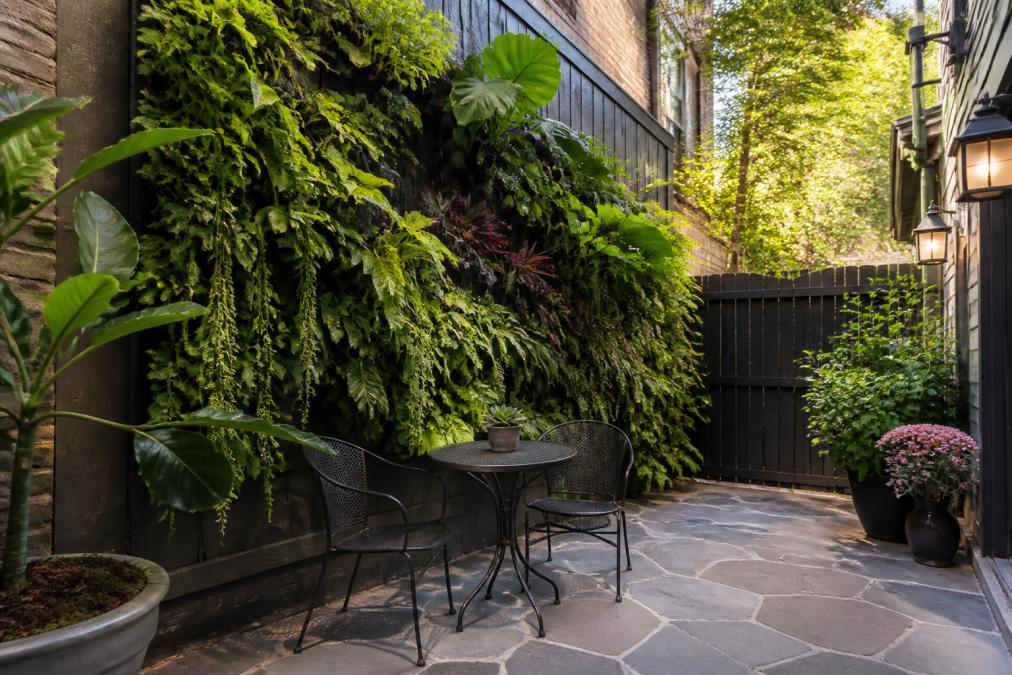 A narrow townhouse backyard featuring a lush vertical green wall planter filled with tropical foliage, with a compact bistro seating set below on a stone patio in warm afternoon light