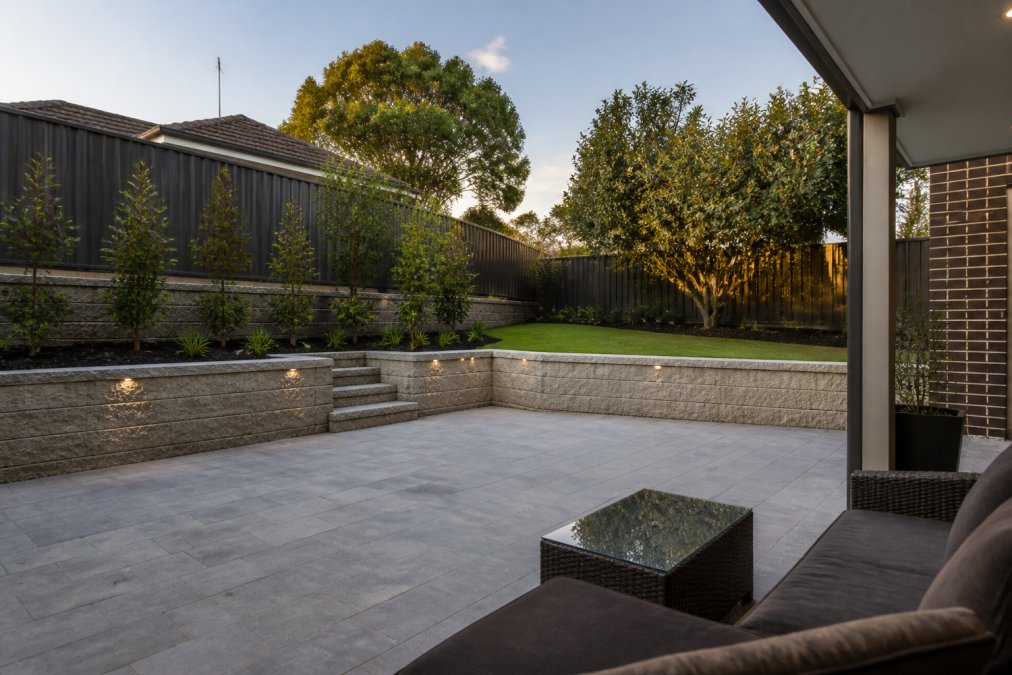 Completed residential backyard with professionally laid paved patio, established retaining wall, and manicured garden beds showcasing finished hardscaping work under natural daylight