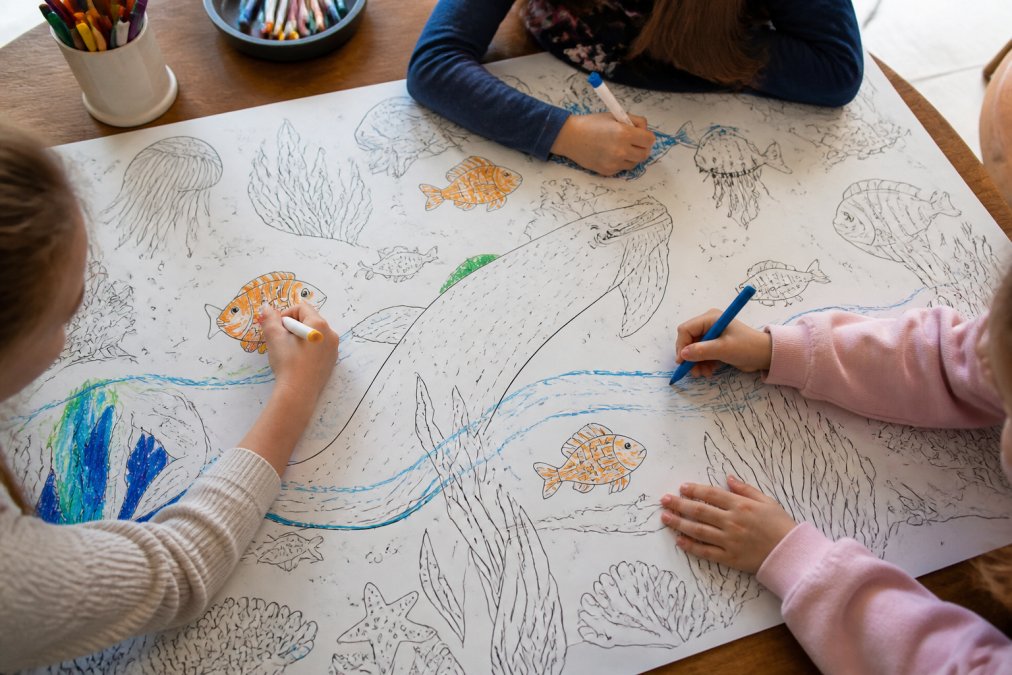 Three children coloring together on large coloring posters spread across a sunlit wooden kitchen table
