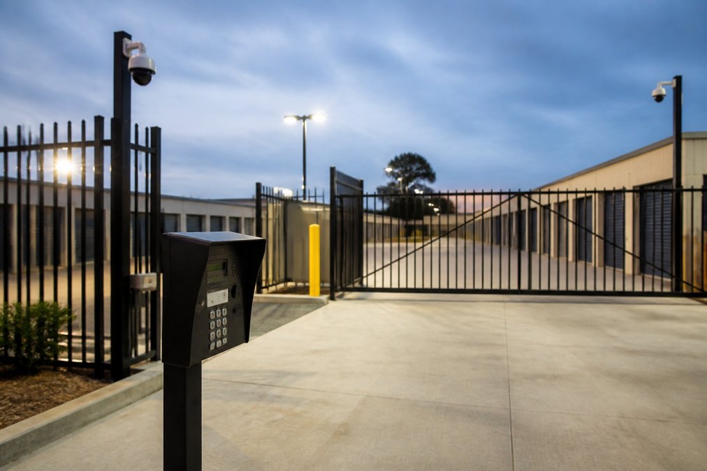 Modern secure storage facility entrance with electronic gate and PIN keypad, CCTV cameras mounted on corner posts, gated perimeter fence, and well-lit access area at dusk