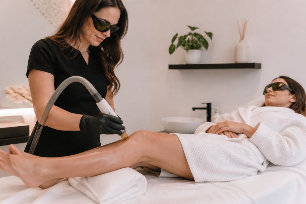 A practitioner uses a cooling laser hair removal device on a client’s leg in a calm, professional clinic setting.