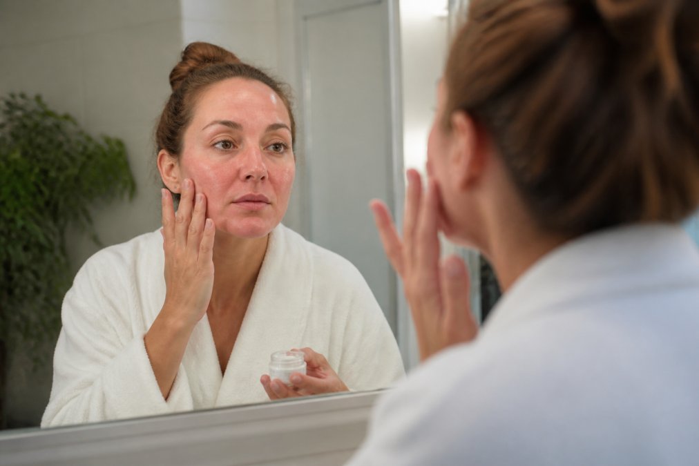 Woman examining her face in a mirror during the first week of recovery, showing mild redness and swelling while gently applying soothing recovery cream with fingertips in soft bathroom lighting