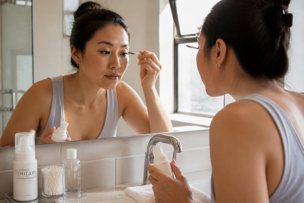 Woman gently cleansing her lash extensions with a spoolie brush and lash shampoo at a bathroom sink, morning light streaming through the window