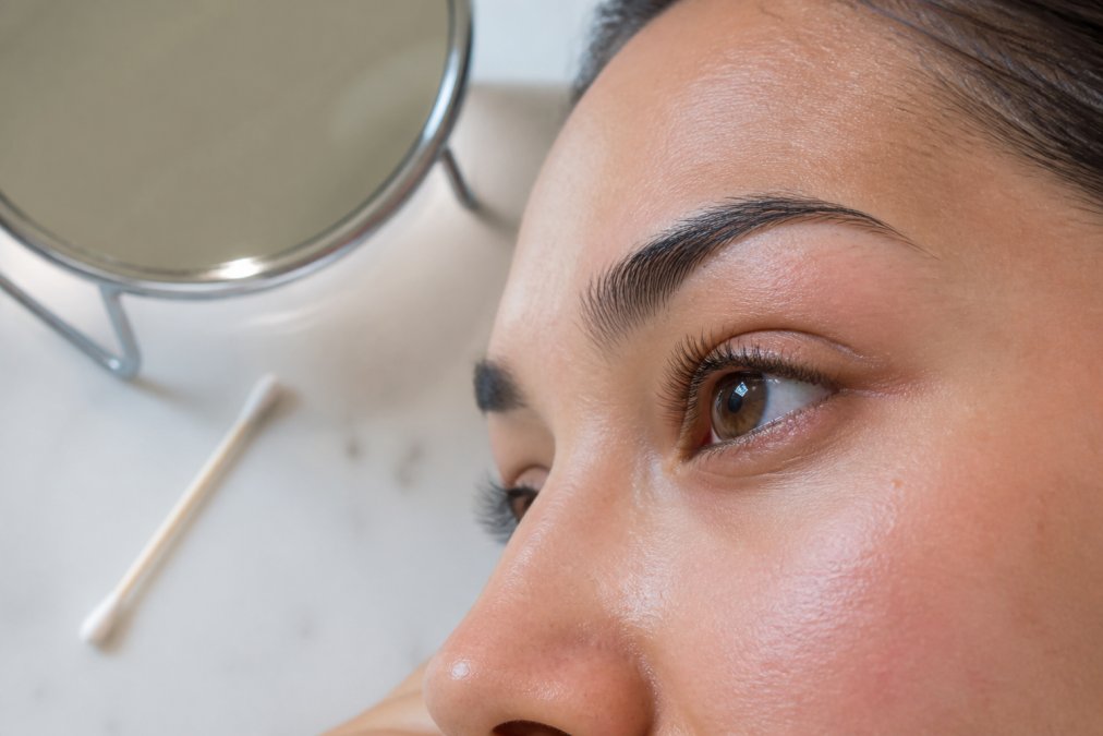 Close-up of a woman's eye area showing natural lashes and a clean lash line, with a cotton swab and small mirror on a clean white countertop