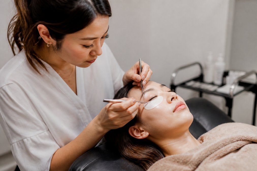 A client reclines in a plush salon chair with eyes closed while a lash artist in white carefully applies extensions with fine tweezers under bright, clean lighting