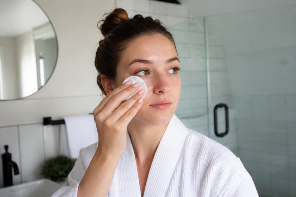 Woman with lifted lashes gently applying oil-free cleanser around her eye area with cotton pad, demonstrating proper aftercare routine in bright bathroom setting