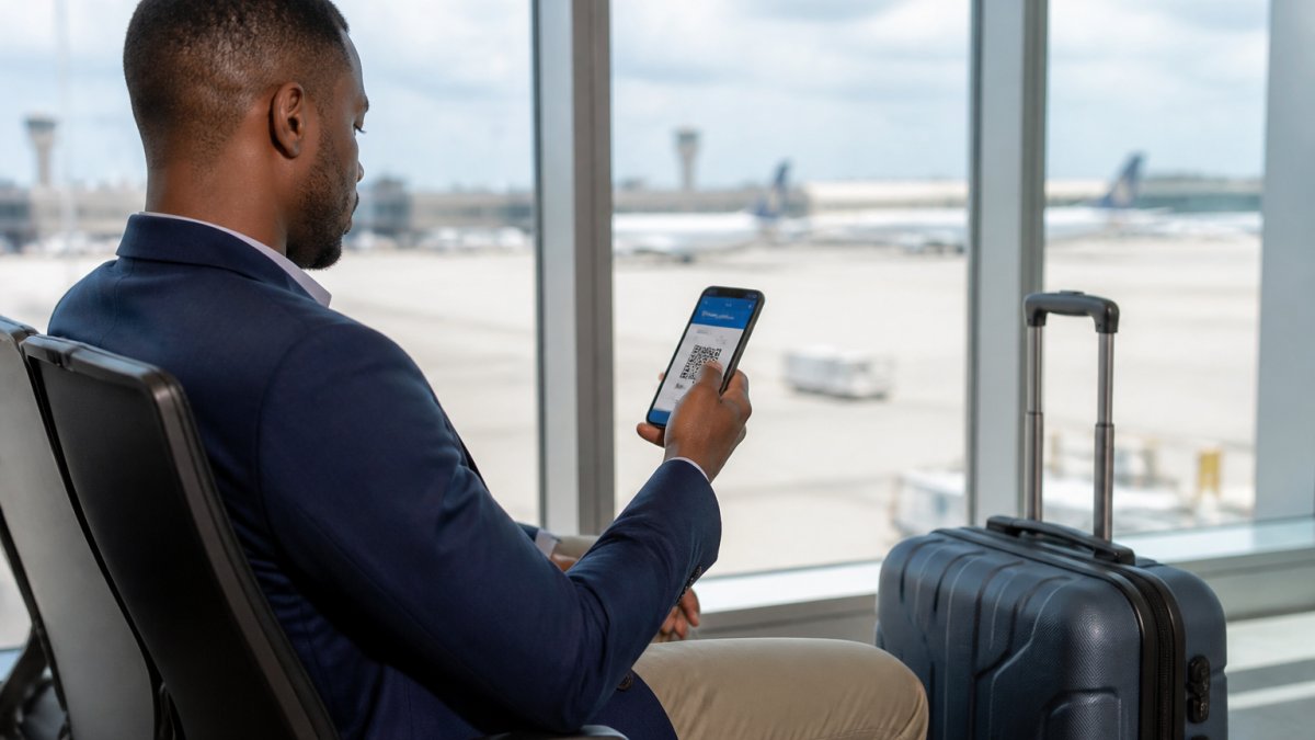 A traveller at an airport departure gate looking at a smartphone showing a boarding pass, suitcase beside them, bright daylight through terminal windows