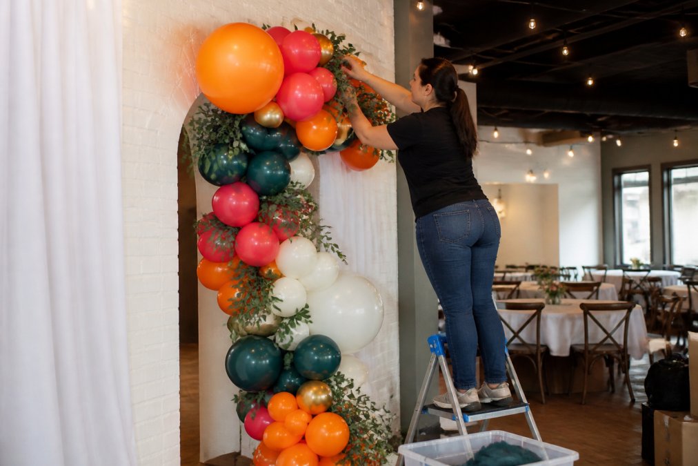 A professional balloon artist installing a colorful organic balloon garland at an event venue, standing on a step ladder with supplies nearby