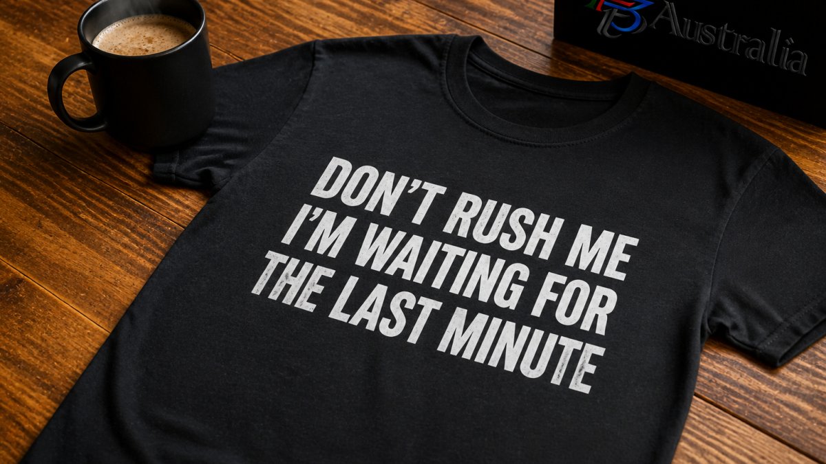 A black cotton t-shirt with the slogan 'Don't Rush Me I'm Waiting For The Last Minute' printed across the chest, laid flat on a timber table beside a steaming coffee mug