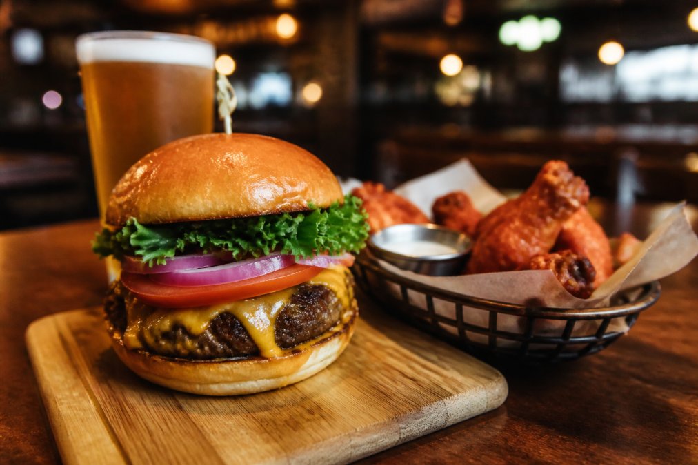 A close-up of a juicy hand-pattied burger with melted cheese and fresh toppings on a wooden board, next to a basket of golden wings and a cold beer, in a warmly lit sports bar setting with big screens softly blurred in the background