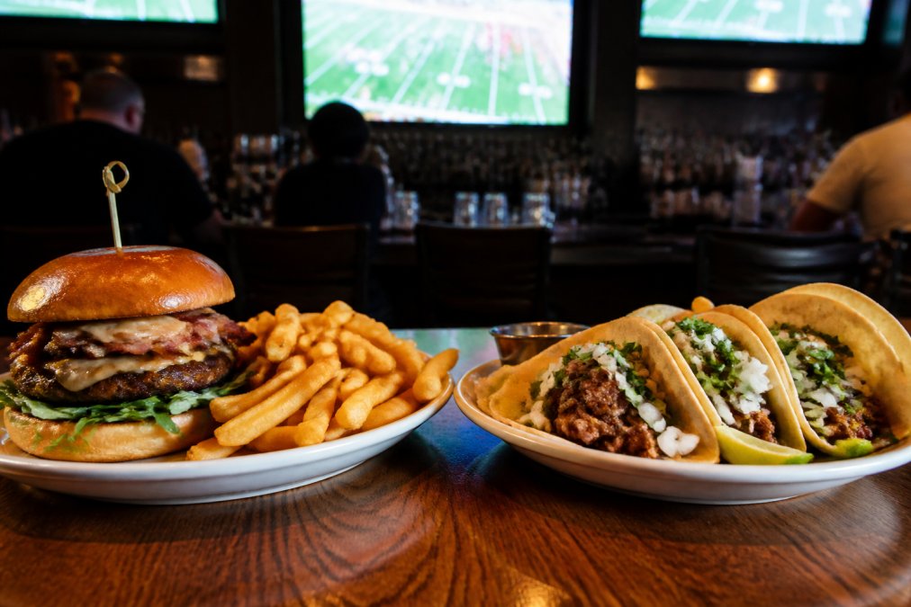 A juicy gourmet burger with fries on one side of a wooden table and a plate of authentic street tacos on the other, set in a dimly lit sports bar atmosphere with a glowing TV screen in the background