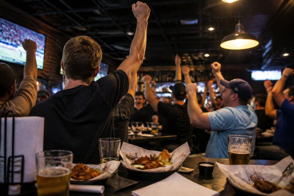 A lively sports bar interior with multiple big screens showing a game, patrons cheering with wings and beer on tables, warm ambient lighting