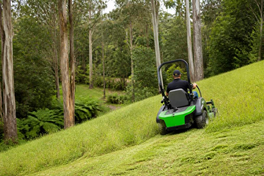 A ride-on mower cutting long grass on a steep, sloping acreage block with tall gum trees and a fern gully in the background