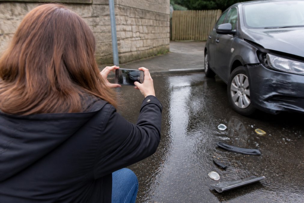 Person taking photographs of accident scene damage with smartphone, documenting evidence on wet pavement with vehicle visible in background