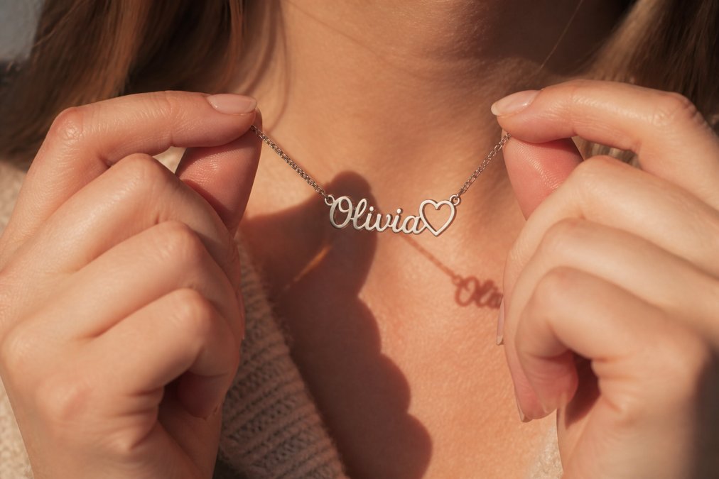 Close-up of a woman's hands holding a personalized name necklace with a heart pendant, polished stainless steel catching warm natural light against a soft neutral background
