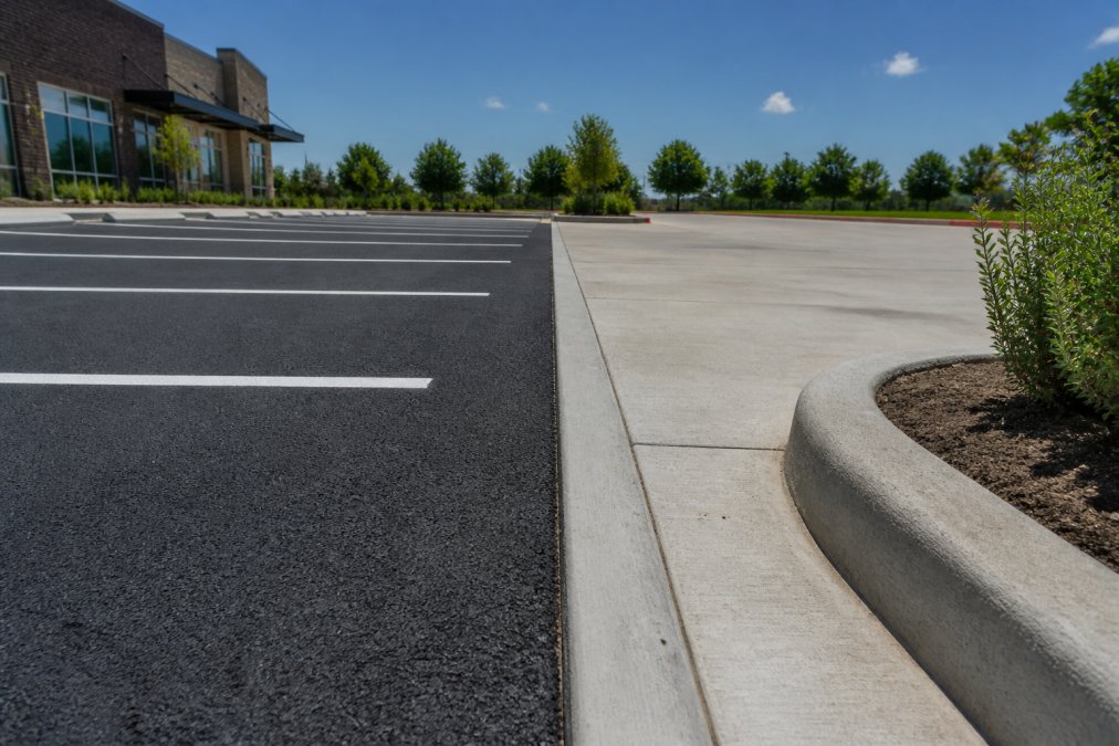 A commercial parking lot with fresh, dark asphalt on one side and a concrete driveway with new curbing on the other, viewed from a low angle under bright Texas sun